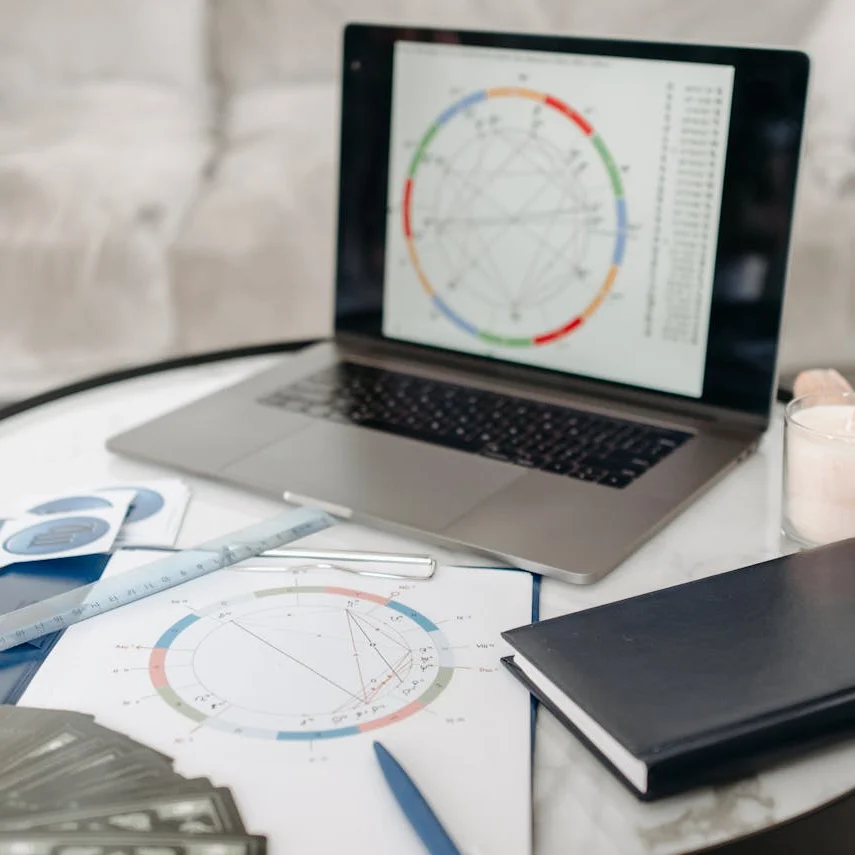 Laptop mit Horoskop, Stift, Lineal, ausgedrucktem Horoskop auf einem Klemmbrett, Tarotkarten, Sternzeichen und Notizbuch auf einem Tisch.